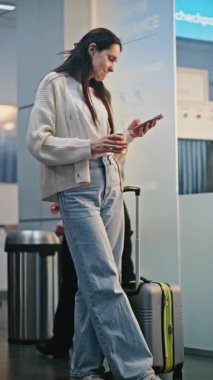 Yoğun Havaalanı Terminali: Akıllı Telefon Kullanan, Kahve İçen Kadın. Anne ve Kızı, Havalimanı Hub Lounge 'da Uçuşu Bekliyor, Aile Tatile Çıkıyor. Yürüyen çeşitli yolcular. Dikey Çekim.