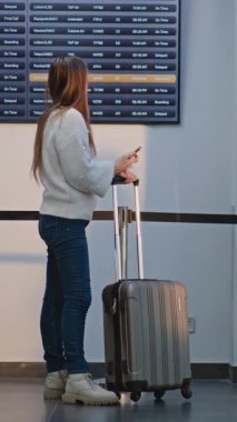 Kalabalık Havaalanı Terminali: Akıllı Telefon Kullanan Kadın, Havayolu Merkezi Biniş Salonu 'nda Uçuşu Bekliyor, Gelen ve Giden Bilgiler Kurulu' nu kontrol ediyor. Çeşitli İnsanlar Yürüyor. Dikey Çekim.