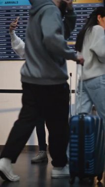 Kalabalık Havaalanı Terminali: Telefonla konuşan bavullu kadın Video Çağrı veya Kayıt Video İçeriği. Gezgin Tatile Çıkıyor. Çeşitli İnsanlar ve Havalimanı Personeli Yürüyüşü. Dikey Çekim.