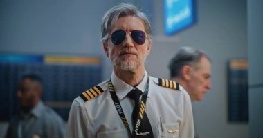 Gözlüklü ve Kameraya Bakan Üniformalı Profesyonel Havayolu Pilotu Portresi. Kalabalık Uluslararası Havalimanı Terminalinde Olgun Uçuş Ekibi Üyesi ve Arka planda Yolcular. Ortam Kapalı