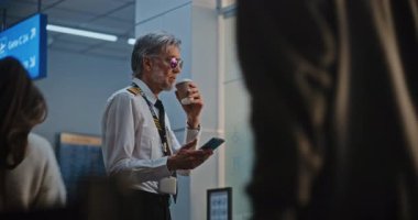 Üniformalı Kılavuz Kahve İçiyor, Telefon Kullanıyor, Ticari Uçuş Bekliyor. Bir kadın havaalanına geliyor Kaptan, Smartphone 'da selfie çekiyor. Kalabalık Uluslararası Havaalanı Terminali. Yavaş Hareket.