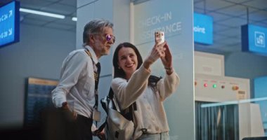 Kalabalık Uluslararası Havaalanı Terminali 'nde Smartphone' da Pilotla Selfie Çeken Kadın Gezgin. Üniformalı Başkomiser kahve içiyor, telefon kullanıyor, ticari uçuşu bekliyor. Yavaş Hareket.