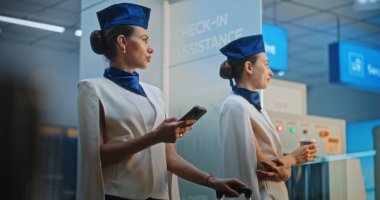 Zarif üniformalı iki güzel uçuş görevlisi konuşuyor, ticari uçuşu bekliyor. Bir kadın akıllı telefon kullanıyor, diğeri kahve içiyor. Kalabalık Uluslararası Havaalanı Terminali. Orta Çekim.