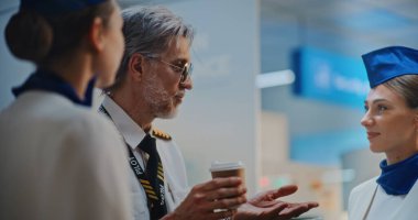 Gözlüklü Hava Yolları Kaptanı Üniformalı İki Güzel Hostesle Konuşuyor, Kahve İçiyor, Ticari Uçuşu Bekliyor. Kalabalık Uluslararası Havaalanı Terminali Uçuş Ekibi. Yakın çekim. Yavaş Hareket