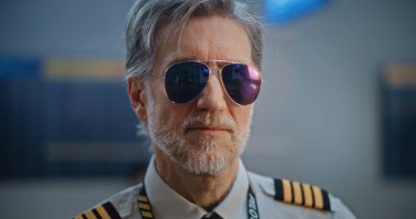 Gözlüklü ve Kameraya Bakan Üniformalı Profesyonel Havayolu Pilotu Portresi. Kalabalık Uluslararası Havalimanı Terminali 'nde Olgun Uçuş Ekibi Üyesi ve Arka planda Yolcular. Kapat..
