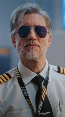 Gözlüklü ve Kameraya Bakan Üniformalı Profesyonel Havayolu Pilotu Portresi. Uluslararası Havalimanı Terminali 'nde Uçuş Ekibi Üyesi ve Arka planda Yolcular. Orta derecede yakın çekim. Dikey Çekim