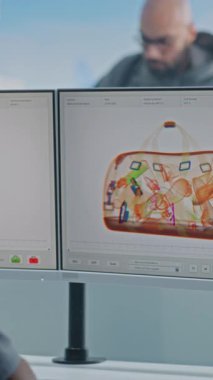 Havaalanı Güvenlik Kontrol Noktası: Afro-Amerikan Güvenlik Subayı Bilgisayar ekranlarında röntgen taraması yapıyor. Tarama işlemi sırasında taşıdığı bagajları olan çeşitli insanlar. Dikey Çekim.