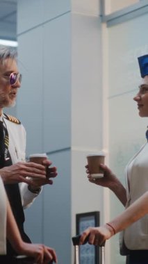 Havayolu pilotu ve üniformalı iki güzel uçuş görevlisi bavulla konuşuyor, kahve içiyor, ticari uçuşu bekliyor. Kalabalık Havaalanı Terminali Uçuş Ekibi. Ağır çekim. Dikey Çekim.