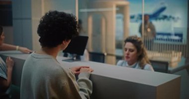 Uluslararası Havalimanı Terminali 'ndeki havalimanı kontrol gişesi: Genç adamla konuşan kadın havayolu işçisi, Pasaport kontrolü sırasında sorgulanıyor. Arkaplanda Güvenlik Kontrol Noktası.