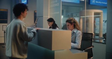 Uluslararası Havalimanı Terminali: Genç Erkek Yolcu Bavulu, Uçuş öncesi Bagaj Bildirimi için Tezgahı yakınında, Bayan Havayolu Ajanıyla Konuşuyor. Arkaplanda Güvenlik Kontrol Noktası.