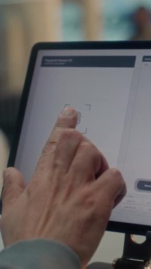 Havalimanı check-in sayacı: Parmak izi taraması için Parmak İzi Tableti Dokunma Ekranı 'na Parmak İzi Koyan Adamın Kapanışı. Havayolu Ajanı Yolcu Biyometrik Verilerini Kontrol Ediyor. Havaalanı Terminali. Dikey Çekim.
