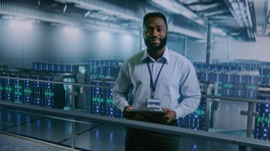 Büyük Veri Merkezi: Afro-Amerikan Tablet Bilgisayar Uzmanı Depo 'da, Kameraya Bakıyor. Bulut Hesaplama Teknolojisi için Sunucu Rafları. Bilgi Dijitalleştirme Konsepti. Orta Çekim.