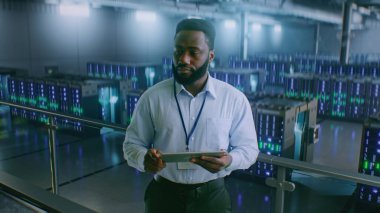 Sunucu Raflı Büyük Veri Merkezi: Profesyonel Afro-Amerikan Uzmanı Tablet Bilgisayar Kullanıyor, Sunucuları Etkinleştiriyor, Depo 'da Duruyor. Bilgi Teknolojisi ve Siber Güvenlik. Orta Çekim.