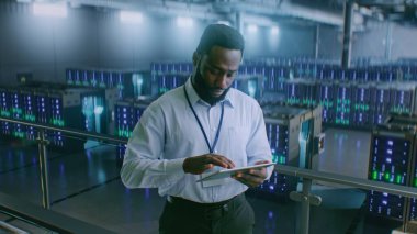 Sunucu Raflı Büyük Veri Merkezi: Profesyonel Afro-Amerikan Uzmanı Tablet Bilgisayar Kullanıyor, Sunucuları Etkinleştiriyor, Depo 'da Duruyor. Bilgi Teknolojisi ve Siber Güvenlik. Orta Çekim.