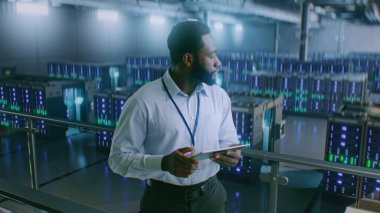 Sunucu Raflı Büyük Veri Merkezi: Profesyonel Afro-Amerikan Uzmanı Tablet Bilgisayar Kullanıyor, Sunucuları Etkinleştiriyor, Depo 'da Duruyor. Bilgi Teknolojisi ve Siber Güvenlik. Orta Çekim.