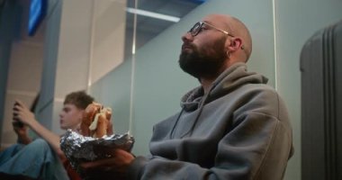 Kalabalık Uluslararası Havaalanı Terminali: Adam Fast Food Burger yerken Havayolu İnbotu 'nun Yatılı Salonu' nda Bavul İçinde Oturuyor. Çeşitli yolcular rötarlı uçuş bekliyor. El bilgisayarı.