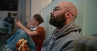 Kalabalık Uluslararası Havaalanı Terminali: İnsan Yiyen Burger 'in Yakın Çekimi, Havayolu Merkezinin Kalkış Salonu' nda Yerde Oturuyor. Çeşitli İnsanlar ve Yolcular Gecikmiş Uçak Uçuşunu Bekliyor.