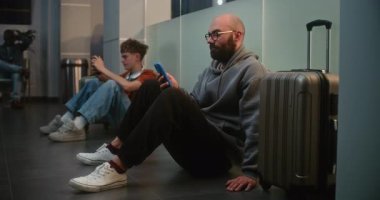 Kalabalık Uluslararası Havaalanı Terminali 'ndeki Çeşitli Yolcular: Bavullu Yorgun Adam Akıllı Telefon, Havayolu Merkezinin Kalkış Salonunda Oturuyor, Gecikmeli Uçuşu Bekliyor.