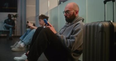Kalabalık Uluslararası Havalimanı Terminali 'ndeki Çeşitli İnsanlar ve Yolcular: Havayolu Hub' un Yatılı Salonunda Bavullu Yorgun Adam, Telefonu kullanarak Gecikmiş Uçak Uçuşunu Bekliyor