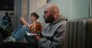 Kalabalık Uluslararası Havalimanı Terminali 'ndeki Çeşitli Yolcular: Adam Fast Food Burger yerken Havayolu İnbotu' nun Kalkış Salonunda Bavullu Oturuyor ve Gecikmeli Uçuşu Bekliyor.