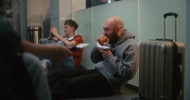 Kalabalık Uluslararası Havalimanı Terminali 'ndeki Çeşitli İnsanlar ve Yolcular: Burger Yiyen Adam, Uçak İnbotu Biniş Salonunda Bavulla Oturan ve Gecikmiş Uçak Uçuşunu Bekleyen Adam.