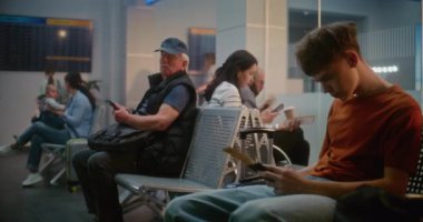 Kalabalık Uluslararası Havaalanı Terminali: Çeşitli Yolcuların ve Yolcuların Yolculuk Salonu 'nda Doli Çekimi. Genç Çocuk Akıllı Telefon Kullanıyor ve Kaydırıyor, Uçak Uçuşunu Bekliyor.