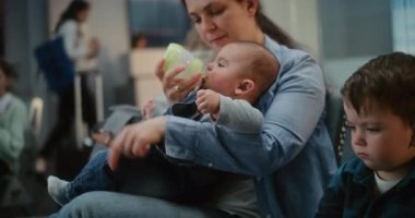 Kalabalık Uluslararası Havaalanı Terminali: Uçak Uçuşunu Bekleyen İki Küçük Çocuğu Olan Anne, Şişeden Bebeği Besleyen Çocuk. Farklı Yolcular ve Yolcular Hava Yolları Merkezinin Kalkış Salonunda.