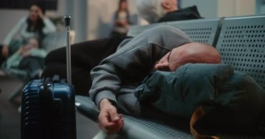 Kalabalık Uluslararası Havalimanı Terminali: Bavullu Yorgun Adam ve Sırt Sırt Çantası Havayolu İnbotu 'nun Yatılı Salonu' nda Uyuyor. Çeşitli yolcular rötarlı uçuş bekliyor. Turizm ve Seyahat.