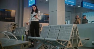 Kalabalık Havaalanı Terminali: Bavullu Mutlu Kadın Güvenlik Kontrolünden Sonra Biniş Salonunda Oturuyor, Uçak Biletleri ve Belgelerini Kontrol Ediyor, Uçuşu Bekliyor. Pasaport Kontrolünde duran yolcular.