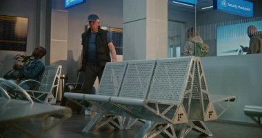 Kalabalık Havaalanı Terminali: Bavulu ve pasaportu olan yaşlı adam güvenlik kontrolünden sonra kalkış salonunda oturuyor, uçak biletini kontrol ediyor, uçuş için bekliyor. Belge Kontrolünde Duran İnsanların Kuyruğu.