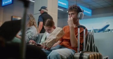 Kalabalık Uluslararası Havaalanı Terminali: Erkek ve Kız, Genç Kardeşler Abur cubur yiyor, Akıllı telefon kullanıyor, Seyahat İçin Uçuşu Bekliyor. Farklı Yolcular Hava Yolları Merkezinin Kalkış Salonunda Oturuyorlar.