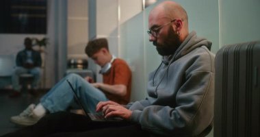 Kalabalık Uluslararası Havalimanı Terminali: Laptop 'ta çalışan adam, e-ticaret e-postaları yolluyor, Havayolu Merkezinin Kalkış Salonunda Yerde Oturuyor. Çeşitli Yolcular Gecikmeli Uçuş Bekliyor.