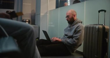 Kalabalık Uluslararası Havaalanı Terminali: Adam Laptop 'ta Uzaktan Çalışırken Havayolu İnbotu Yatılı Salonu' nda Bavulla Oturuyor. Çeşitli İnsanlar Gecikmiş Uçak Uçuşunu Bekliyor.