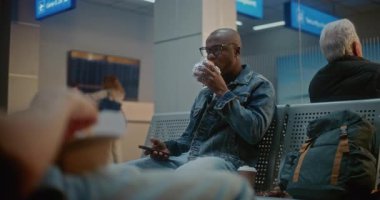Kalabalık Uluslararası Havalimanı Terminali: Afro-Amerikan Adam Fast Food yiyor ve akıllı telefon kullanıyor. Çeşitli Yolcular Gecikmiş Uçak Uçuşunu Bekliyor.