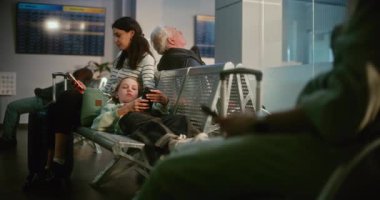 Kalabalık Havalimanı Terminalinde Anne ve Kızı: Koltuklarda uzanan Bitkin Ergen Kız, Telefonu kullanan kadın. Çeşitli İnsanlar ve Yolcular Hava Yolları Merkezinin Kalkış Salonunda Gecikmeli Uçuş Bekliyor.