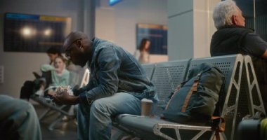 Kalabalık Uluslararası Havaalanı Terminali: Afro-Amerikan Adam Burger Yiyor ve Kaydırma Telefonu Gecikmeli Uçuşu Bekliyor. Farklı İnsanlar ve Yolcular Hava Yolları Merkezinin Kalkış Salonunda Oturuyorlar.