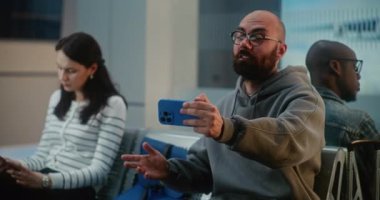 Kalabalık Uluslararası Havaalanı Terminali: Video Sohbeti 'nden Adam Konuşuyor, Trip' ten Video İçeriğini Çekiyor Akıllı Telefon Kullanıyor, Uçuşu Bekliyor. Farklı Yolcular Hava Yolları Merkezinin Kalkış Salonunda Oturuyorlar.