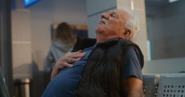 Kalabalık Uluslararası Havaalanı Terminali: Yaşlı Adam Göğsünü Tutuyor, Kalp krizi geçiriyor ya da Hava Yolları İnbotu Salonunda Enfarktüs geçiriyor. Çeşitli İnsanlar ve Havalimanı Personeli Kıdemli Yolcuya Yardım Ediyor.