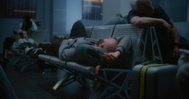 Kalabalık Uluslararası Havaalanı Terminali: Yorgun Adam Gece Havalimanı 'nın Kalkış Salonunda Sırt çantasında Uyuyor. Çeşitli Yolcular ve Gezginler Gecikmiş Uçak Uçuşunu Bekliyor.