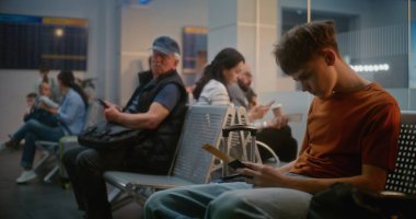 Kalabalık Uluslararası Havaalanı Terminali: Çeşitli Yolcuların ve Yolcuların Yolculuk Salonu 'nda Doli Çekimi. Genç Çocuk Akıllı Telefon Kullanıyor ve Kaydırıyor, Uçak Uçuşunu Bekliyor.