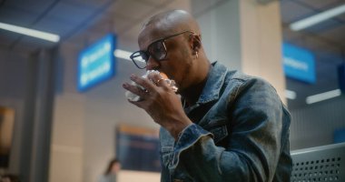 Kalabalık Uluslararası Havalimanı Terminali: Afro-Amerikan Adam Burger Yiyor ve Akıllı Telefon Kullanıyor Havayolu Merkezinin Yatılı Salonunda Oturuyor. Gecikmeli uçuş bekleyen yolcu. Ortalama Yakın Çekim.