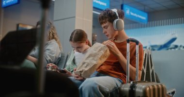 Kalabalık Uluslararası Havaalanı Terminali: Erkek ve Kız, Genç Kardeşler Abur cubur yiyor, Akıllı telefon kullanıyor, Seyahat İçin Uçuşu Bekliyor. Farklı Yolcular Hava Yolları Merkezinin Kalkış Salonunda Oturuyorlar.