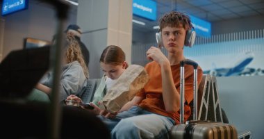 Kalabalık Uluslararası Havaalanı Terminali: Erkek ve Kız, Genç Kardeşler Abur cubur yiyor, Akıllı telefon kullanıyor, Seyahat İçin Uçuşu Bekliyor. Farklı Yolcular Hava Yolları Merkezinin Kalkış Salonunda Oturuyorlar.