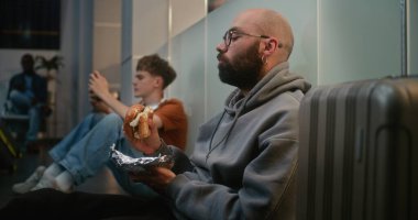 Kalabalık Uluslararası Havalimanı Terminali 'ndeki Çeşitli Yolcular: Adam Fast Food Burger yerken Havayolu İnbotu' nun Kalkış Salonunda Bavullu Oturuyor ve Gecikmeli Uçuşu Bekliyor.