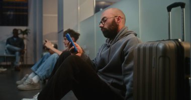 Kalabalık Uluslararası Havalimanı Terminali 'ndeki Çeşitli İnsanlar ve Yolcular: Havayolu Hub' un Yatılı Salonunda Bavullu Yorgun Adam, Telefonu kullanarak Gecikmiş Uçak Uçuşunu Bekliyor