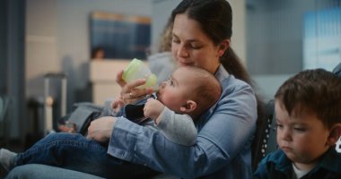 Kalabalık Uluslararası Havaalanı Terminali: Uçak Uçuşunu Bekleyen İki Küçük Çocuğu Olan Anne, Şişeden Bebeği Besleyen Çocuk. Farklı Yolcular ve Yolcular Hava Yolları Merkezinin Kalkış Salonunda.