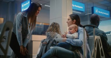 Kalabalık Uluslararası Havaalanı Terminali: Yetişkin Kadın Bebek Oturuyor ve Uçak Bekliyor. Kadın havaalanı işçisi, kadınla konuşuyor. Hava Yolları Merkezinin Ayrılış Salonundaki Çeşitli Yolcular.