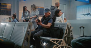 Uluslararası Havaalanı Terminali: Akıllı Telefon Kullanan Yaşlı Adam, Uçak Uçuşunu Bekliyor. Muhtelif Turistler ve Yolcular Kalabalık Havayolu Merkezinin Kalkış Salonunda Oturuyorlar, Seyahate Çıkıyorlar.