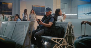 Uluslararası Havaalanı Terminali: Akıllı Telefon Kullanan Yaşlı Adam, Uçak Uçuşunu Bekliyor. Muhtelif Turistler ve Yolcular Kalabalık Havayolu Merkezinin Kalkış Salonunda Oturuyorlar, Seyahate Çıkıyorlar.