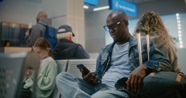 Uluslararası Havaalanı Terminali: Bavullu Afro-Amerikan Adam Akıllı Telefon kullanarak Uçağı Bekliyor. Kalabalık Havayolu Merkezinin Ayrılış Salonundaki Çeşitli İnsanlar ve Yolcular.